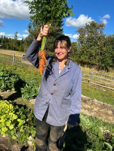 Una mujer sonriente está de pie en un jardín soleado, sosteniendo con orgullo una zanahoria grande y de forma peculiar que acaba de cosechar. Lleva una chaqueta azul y pantalones de trabajo.