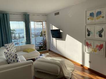 L'image représente un salon serein avec une vue imprenable sur la mer, avec des meubles blancs, des œuvres d'art vibrantes et un téléviseur accroché au mur, dégageant une impression de calme.