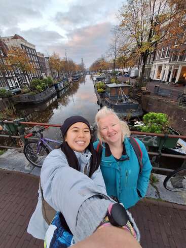 Dos mujeres sonrientes se toman un selfie en un puente sobre un pintoresco canal de Ámsterdam, rodeadas de bicicletas, casas flotantes, árboles otoñales y edificios históricos.