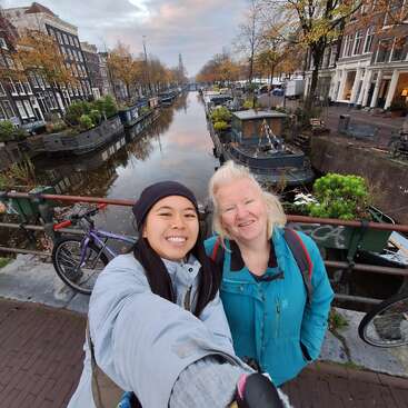 Dos mujeres sonrientes se toman un selfie en un puente sobre un pintoresco canal de Ámsterdam, rodeadas de bicicletas, casas flotantes, árboles otoñales y edificios históricos.