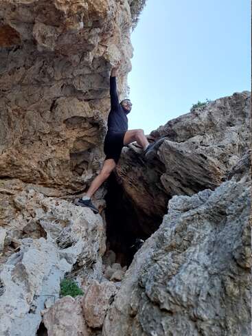 A man in athletic attire is climbing a rocky cliff, with his right arm raised and left leg bent, set against a clear blue sky.