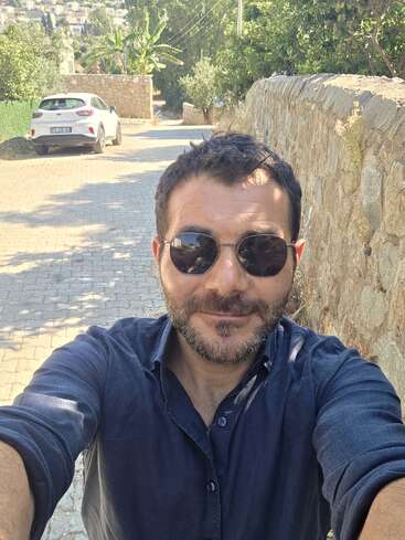 A man with short dark hair and beard, wearing sunglasses and a dark shirt, takes a selfie on a sunny cobblestone street lined with stone walls.