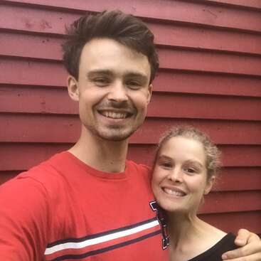 Um casal de jovens sorridentes está bem juntinho, tirando uma selfie na frente de uma parede de madeira vermelha. Os dois parecem felizes, relaxados e estão curtindo a companhia um do outro.