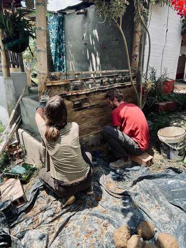 Two people are working on a mud wall, surrounded by plants and tools, in a sunny outdoor setting, with a white building in the background.