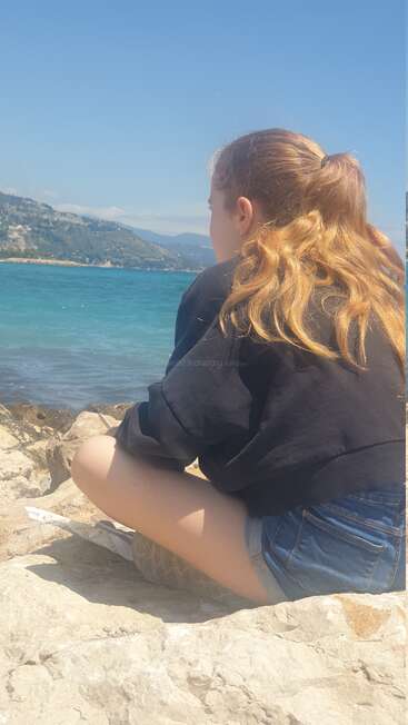 A girl with long blonde hair sits on rocks by the sea, wearing a black sweatshirt and denim shorts, gazing thoughtfully at the beautiful blue water.