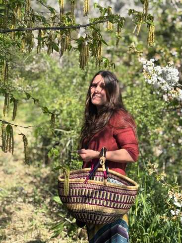 A woman stands outdoors surrounded by lush greenery and flowering branches, smiling. She holds a colorful woven basket and wears a red top and patterned skirt.