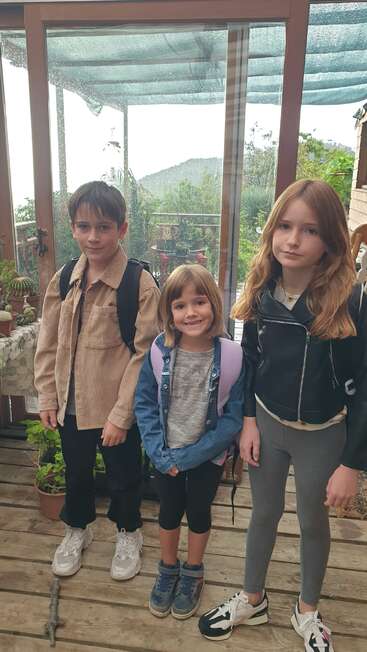 Three children with backpacks stand indoors by a glass door, possibly ready for school. Two look serious, one smiles brightly. Plants decorate the wooden floor.