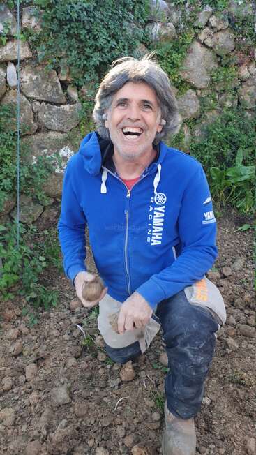 A smiling man kneels on rocky soil, wearing a blue Yamaha Racing jacket and work pants. He holds pieces of dirt, surrounded by greenery and stone wall.