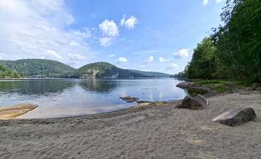 A tranquil lake scene shows calm water, sandy beach, scattered large rocks, and lush green forest. Rolling hills and blue sky with fluffy clouds complete the view.