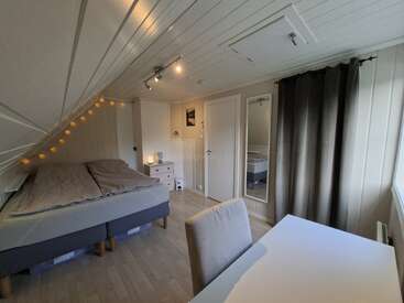 This cozy bedroom has a slanted ceiling, double bed with gray bedding, string lights, desk, chair, dresser, full-length mirror, and large window with dark curtains.