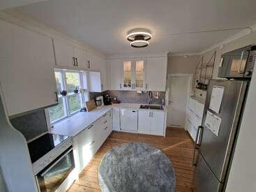 Esta cocina moderna cuenta con armarios blancos, electrodomésticos de acero inoxidable, una alfombra redonda gris, suelo de madera, ventanas grandes, estanterías abiertas y una lámpara de techo de estilo contemporáneo.