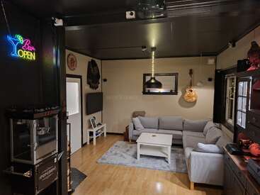 Una acogedora sala de estar cuenta con un sofá modular gris, una mesa de centro blanca, una guitarra colgada en la pared, un letrero de neón que dice «Bar Open», una máquina de palomitas, un televisor y una decoración ecléctica. Iluminación cálida.