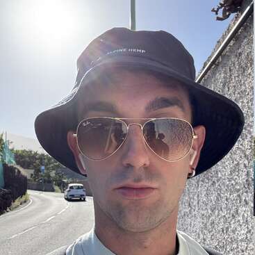 Das Bild zeigt einen Mann, der einen schwarzen Hut, ein weißes Hemd und eine Sonnenbrille trägt und am Straßenrand steht, während hinter ihm ein Auto fährt.