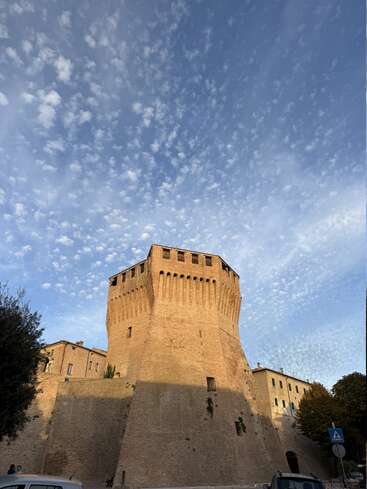 A towering medieval fortress of tan bricks stands under a vibrant blue sky, dotted with scattered clouds, surrounded by trees, cars, and street signs at sunset.