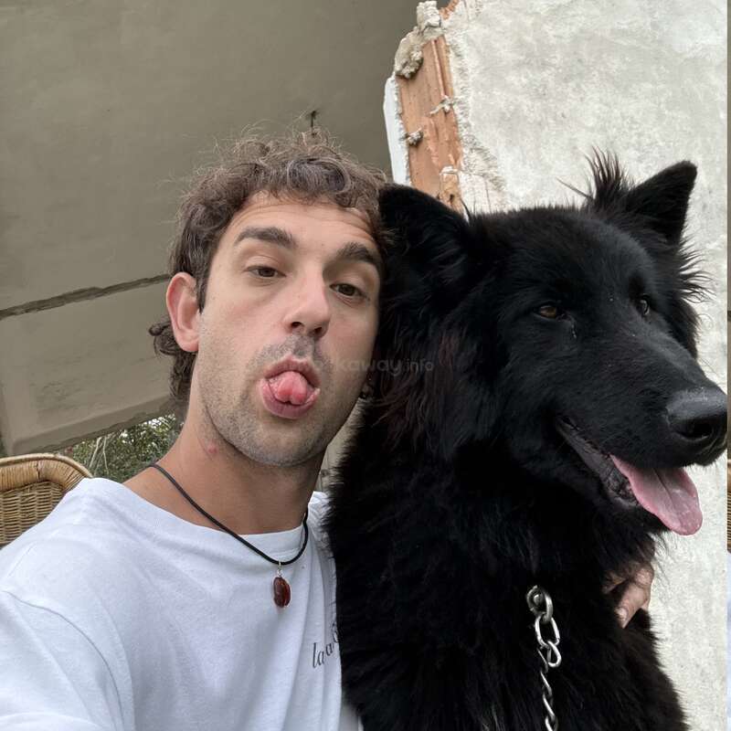 Un joven posa junto a su peludo perro negro; ambos sacan la lengua en tono juguetón. Parecen felices, sentados al aire libre junto a una pared parcialmente dañada.