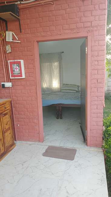 The image shows an open door leading into a small room with a bed, light curtains, a pink brick wall, first aid box, and marble flooring.