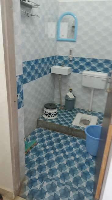 This image shows a small bathroom with blue-patterned tiles, a squat toilet, a blue bucket, a small sink, a mirror, and a trash bin.