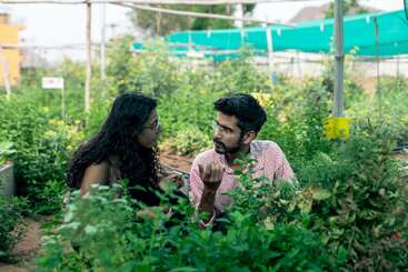 Two people are sitting and talking in a lush, green garden, surrounded by various plants. The environment looks peaceful, and they appear engaged and focused.