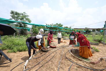 Several women and one man work on a farm, tending to soil and plants. Drip irrigation pipes are visible. The setting appears rural, under clear skies.