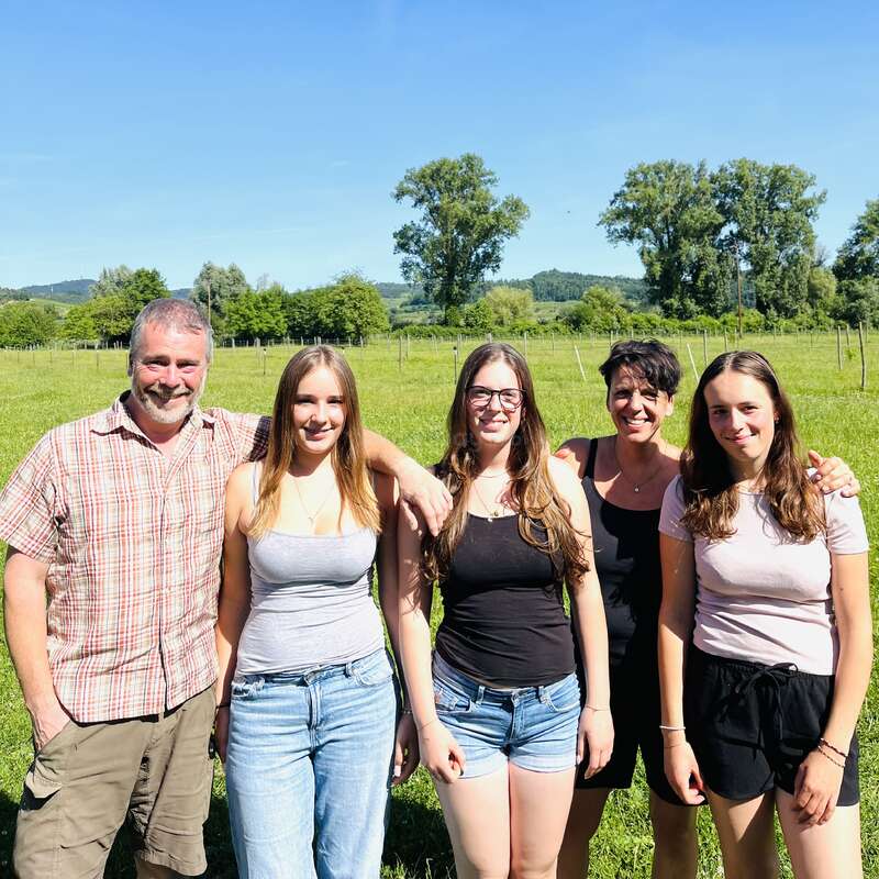 Eine glückliche fünfköpfige Familie steht an einem sonnigen Tag gemeinsam auf einer grünen Wiese, lächelt und hat Bäume und den blauen Himmel im Hintergrund.