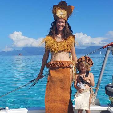 Una mujer y un niño, ambos vestidos con trajes tradicionales de inspiración polinesia, posan en una embarcación con aguas cristalinas y montañas lejanas al fondo.