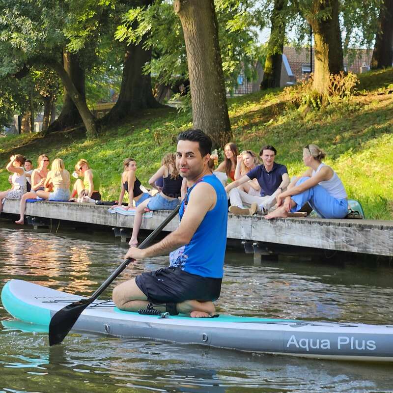 A man paddles a kayak on a calm river, surrounded by lush green trees, while people sit and chat on the riverside dock, enjoying nature.