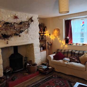 Uma aconchegante sala de estar com lareira de pedra, luzes decorativas, sofá confortável, livros, harpa, cesta, tapete estampado e decorações festivas, que transmitem calor e charme.