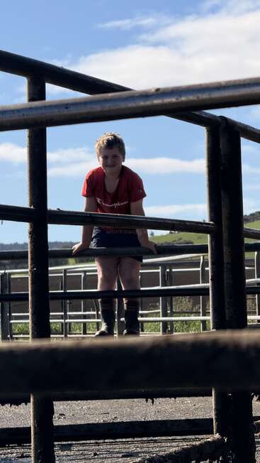 A young boy in a red shirt and boots sits on metal fencing outdoors, smiling, with a blue sky, clouds, and green hills in the background.