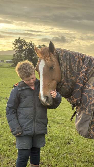 A young boy in a jacket gently caresses a brown horse’s face. They share a quiet, tender moment together in a green field at sunset.