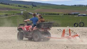 Two kids ride a red ATV across a dusty, gravel area, towing a small orange farming implement. Green fields and rolling hills stretch out behind them.