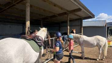 Three children wearing helmets are tending to two white horses under a shaded stable. One child is mounting a saddled horse; the others are preparing the second horse.
