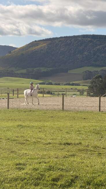 A child in a helmet rides a white horse in a fenced arena, surrounded by lush green fields, hills, and a cloudy sky in the background.