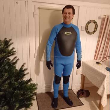 L'image représente un homme debout dans une pièce, vêtu d'une combinaison de plongée bleue avec des accents et des gants noirs, posant pour l'appareil photo devant une porte blanche.