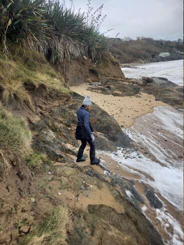 Une personne portant un bonnet gris et une veste bleue marche le long d'une plage rocheuse, avec une falaise et une zone herbeuse à gauche, et une étendue d'eau à droite.