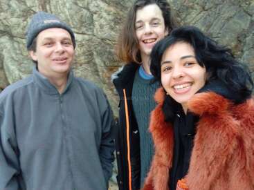 L'image montre trois personnes debout devant une paroi rocheuse, dont deux portent des manteaux et une un chapeau, et qui sourient à l'appareil photo.