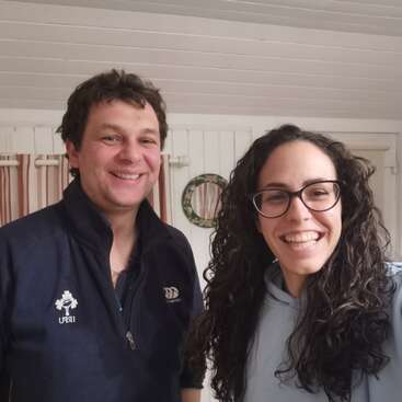 The image shows two people standing side by side, smiling at the camera, with a white wall and ceiling in the background.