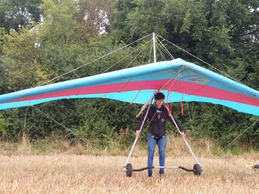 L'image représente un homme debout dans un champ, tenant un deltaplane coloré avec une voile bleue et rose, vêtu d'une veste noire et d'un jean bleu.