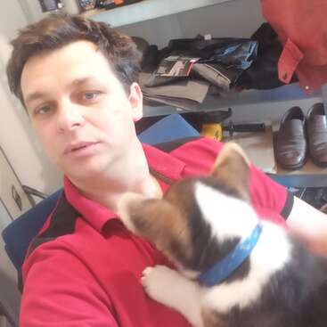 L'image montre un homme en chemise rouge, un petit chien sur l'épaule, assis devant une étagère encombrée de chaussures et d'autres objets.