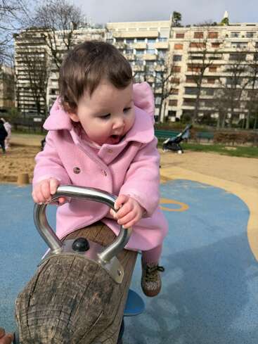 A baby wearing a pink coat enjoys a playground seesaw. She looks excited and curious, gripping the handles tightly, with buildings and trees in the background.