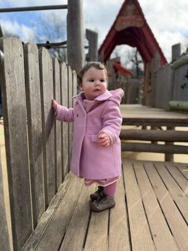A young child in a pink coat stands on a wooden playground structure, holding onto a fence, with a red-roofed playhouse in the background.