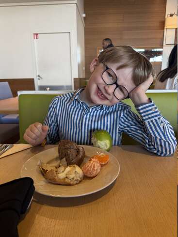 A smiling young boy with glasses, wearing a blue striped shirt, sits at a table with bread, a clementine, and an apple on his plate.