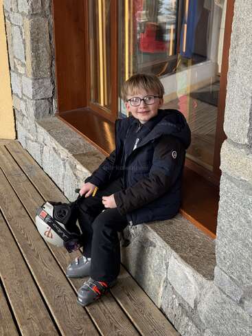 A smiling young boy wearing glasses and a winter jacket sits on a stone bench. He holds a ski helmet, wearing ski boots, ready for adventure.