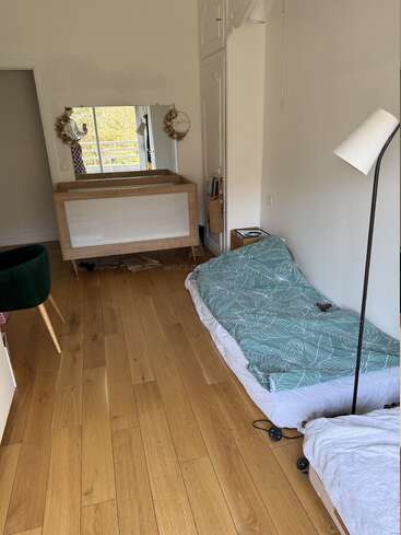 Ein schlichtes, gemütliches Schlafzimmer mit Holzboden, einer Einzelmatratze mit Bettwäsche im grünen Blattmuster, einer Stehlampe, einem Spiegel, einer Kommode, einem grünen Stuhl und minimalistischer Einrichtung.