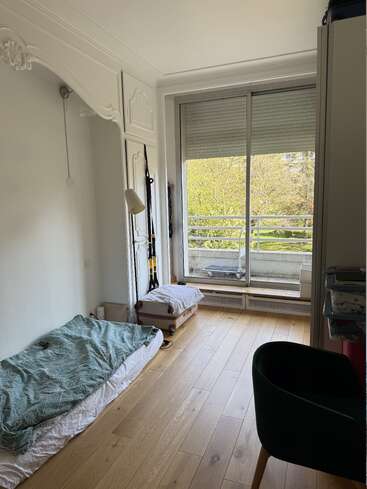 Ein gemütliches, minimalistisch eingerichtetes Schlafzimmer mit einer Matratze auf dem Holzboden, einer grünen Decke, einem großen Fenster mit Blick auf Bäume, weiß verzierten Wänden, einem Koffer und einem schwarzen Stuhl.