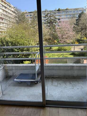 Ein Balkon mit einem kleinen Wagen bietet einen Blick auf einen Garten voller Bäume und Grünpflanzen. Hohe Wohnhäuser umgeben die Gegend unter einem strahlend blauen Himmel.