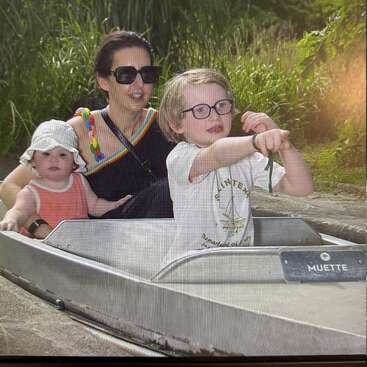 A woman with two children sits in a small boat, surrounded by greenery. The older child points ahead excitedly, while the younger wears a sunhat.
