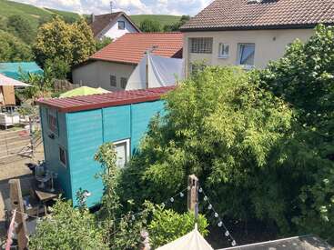 Essa imagem mostra uma casinha azul vibrante com telhado vermelho, cercada por uma vegetação exuberante, casas vizinhas e luzes decorativas, sob um céu ensolarado.