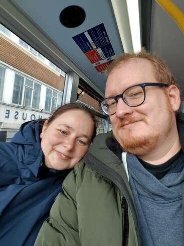 Una pareja sonriente se hace un selfi en primer plano en el interior de una casa, vestida con chaquetas de abrigo. Detrás de ellos se ven unos grandes ventanales y un edificio en el que se lee la palabra «HOUSE». Ambiente acogedor.