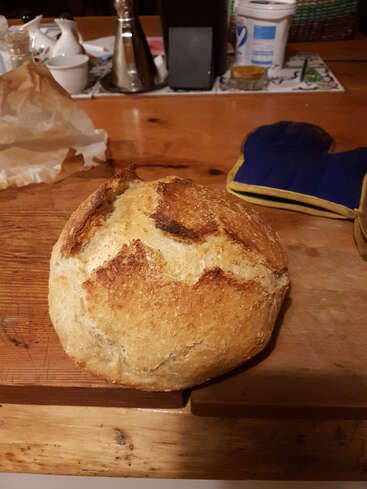Ein runder, rustikaler Laib selbstgebackenes Brot liegt auf einem hölzernen Schneidebrett, während im warm beleuchteten Hintergrund ein blauer Ofenhandschuh und Küchenutensilien zu sehen sind.