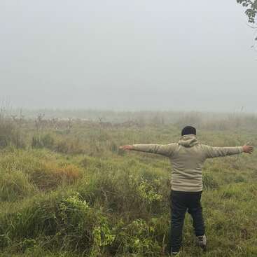Una persona se encuentra en un campo neblinoso con los brazos extendidos, frente a un lejano grupo de ciervos que pastan tranquilamente entre la hierba alta y la naturaleza salvaje.